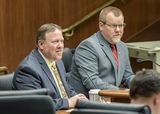 Steve Dennis sits at the testifiers' table and speaks to someone out of view. Representative Jurgens sits next to him.
